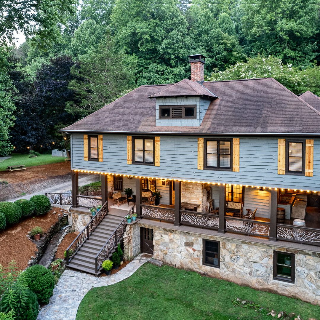 Blue wooden Bed & Breakfast with a stone foundation and wrap-around porch, lit by string lights in a wooded yard. - Clayton, GA