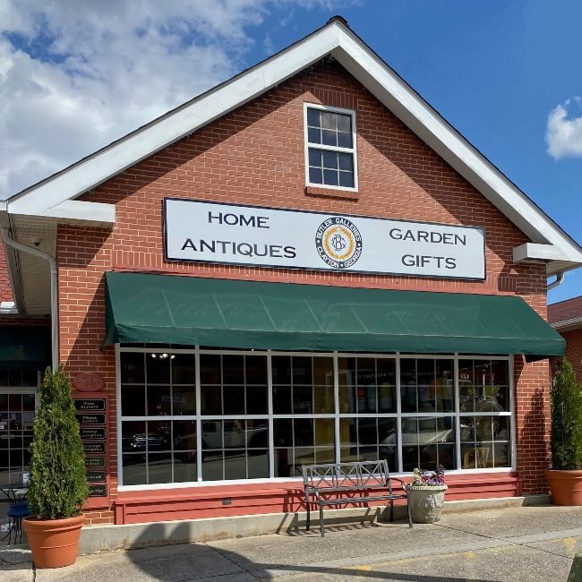 Brick storefront of an antiques shop with a green awning, large front windows, and outdoor bench; sign reads 'Home Antiques Garden Gifts' beneath a gable roof. - Clayton, GA