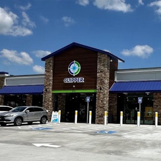 Exterior of a Clipper store with stone pillars, brown wood facade, blue awnings, and parked cars under a sunny sky. - Clayton, GA