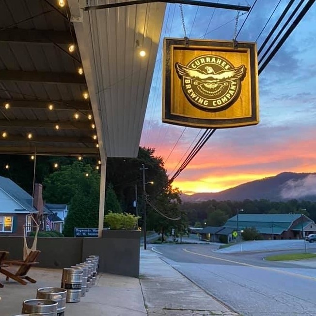 Exterior of Currahee Brewing Company at sunset, with a hanging wooden sign, string lights, and a quiet street nearby. - Clayton, GA