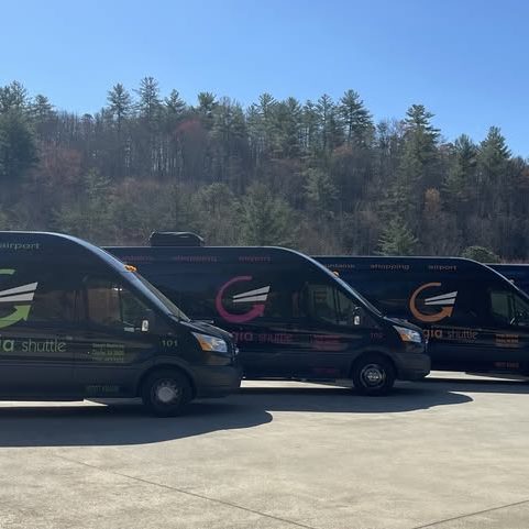 Three dark Georgia Shuttle vans lined up on a paved lot with trees in the background. - Clayton, GA