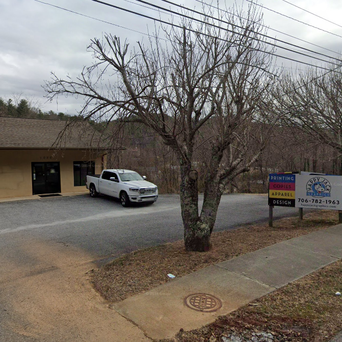A small business storefront with a gravel parking lot, a white pickup truck parked near the entrance, and bare trees in front; a sidewalk runs along the street on the right under a cloudy sky. - Clayton, GA