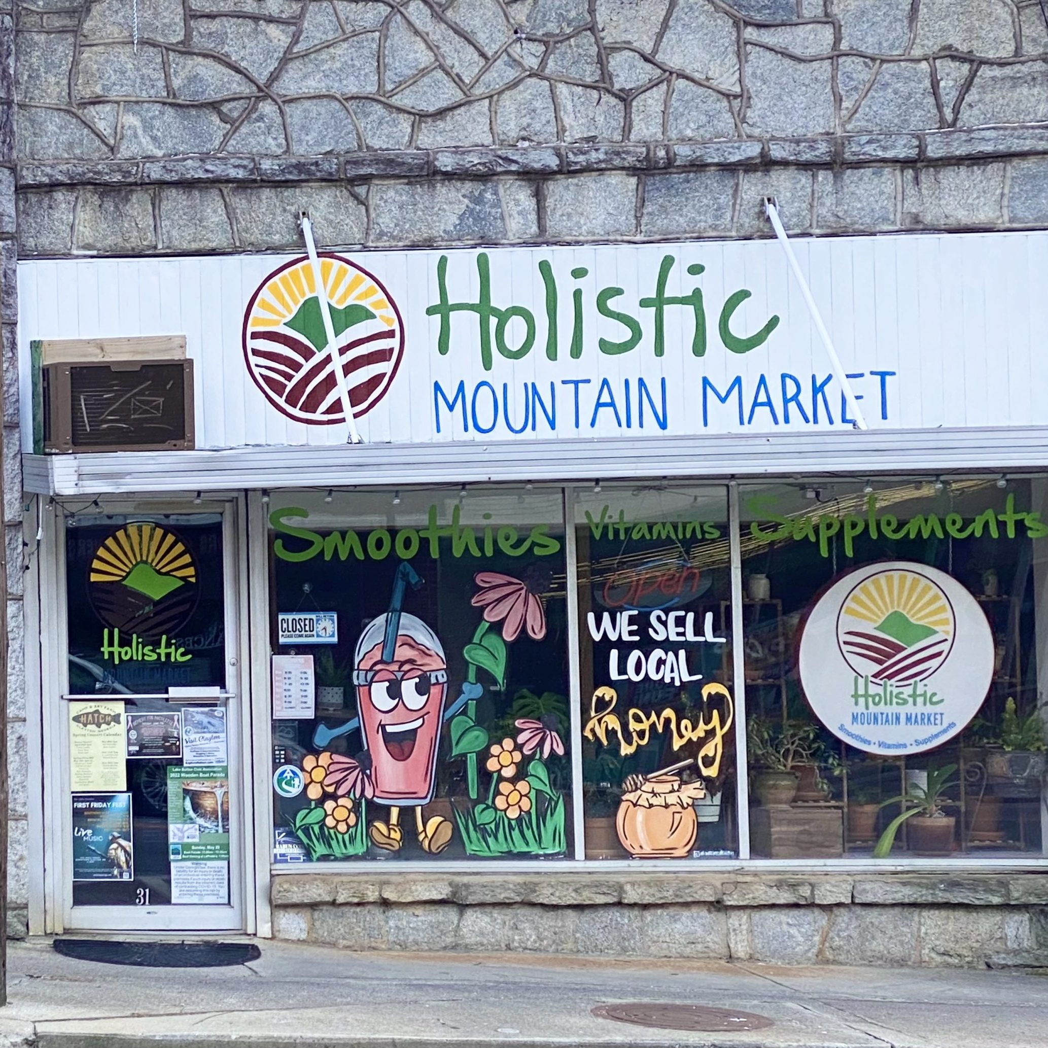 Storefront sign for Holistic Mountain Market with a sun-mountain logo and colorful window art advertising smoothies and vitamins/supplements. - Clayton, GA