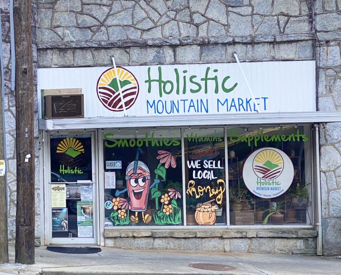Storefront sign for Holistic Mountain Market with a sun-mountain logo and colorful window art advertising smoothies and vitamins/supplements. - Clayton, GA