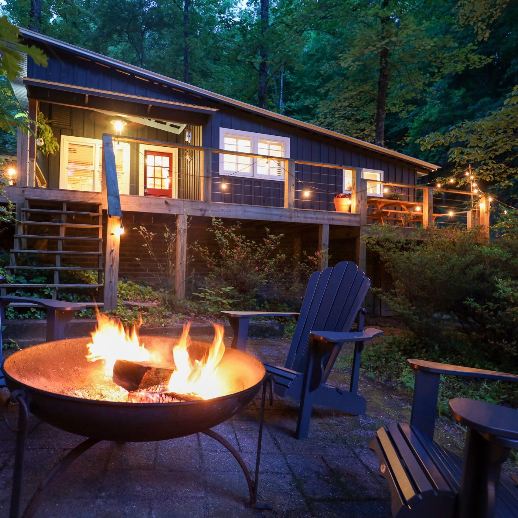 Nighttime backyard with a wooden cabin, string lights, and a glowing fire pit surrounded by blue Adirondack chairs. - Clayton, GA