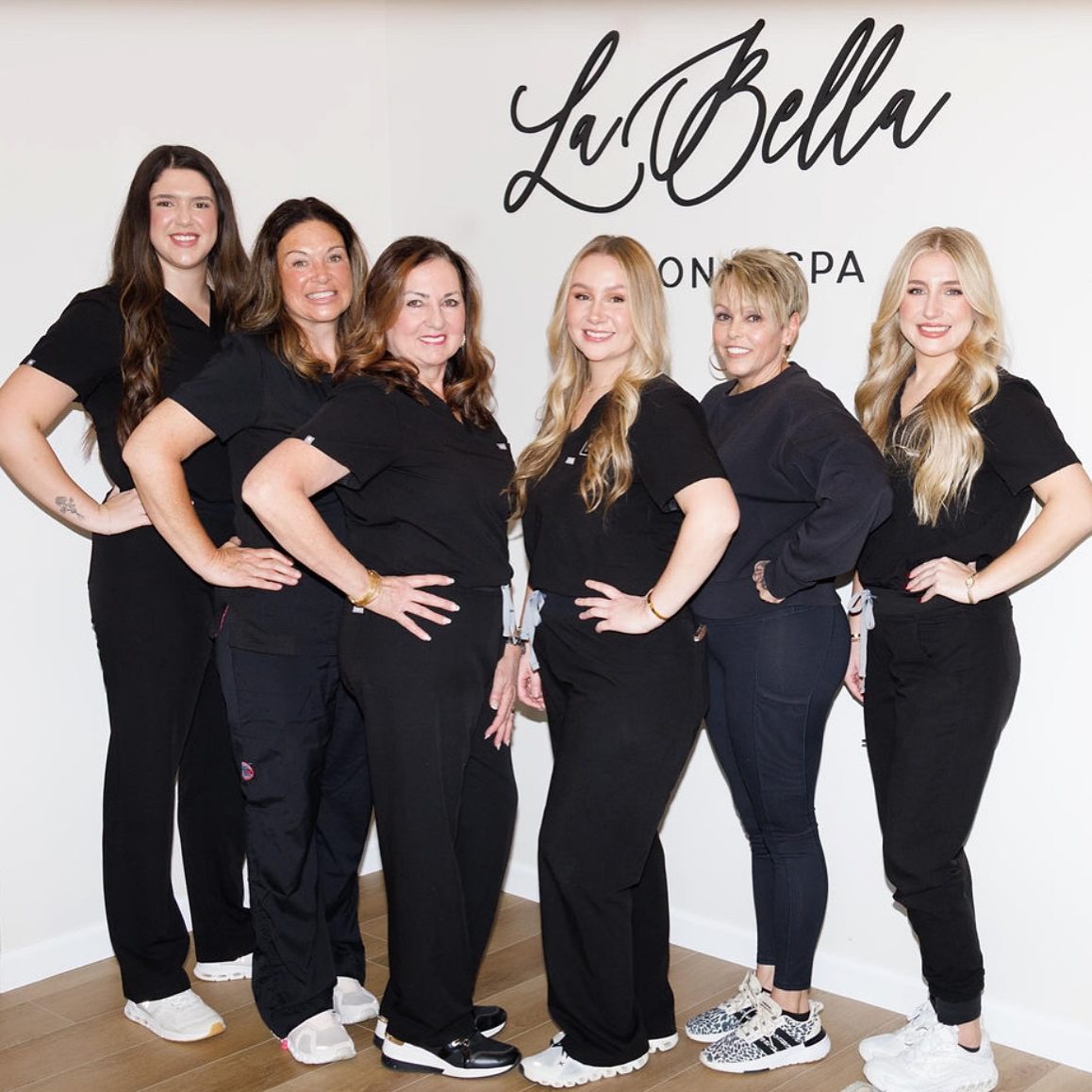 Six women in black scrubs pose together in a spa or salon, with the La Bella logo on the wall behind them. - Clayton, GA