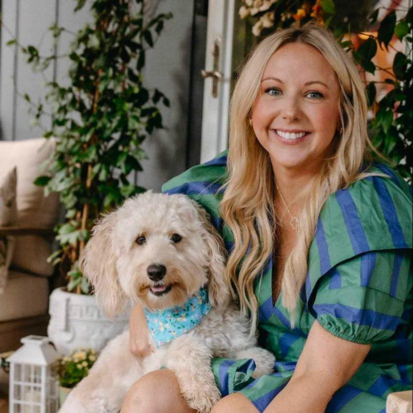 Smiling blonde woman in a green and blue dress sits with a light-colored poodle wearing a blue bandana indoors or in a patio setting. - Clayton, GA