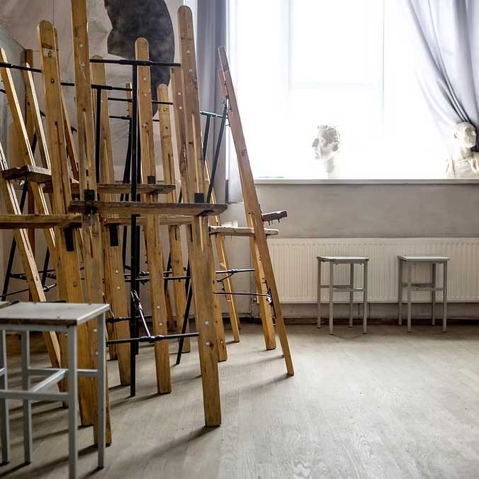 Art studio with tall wooden easels stacked together, metal stools, and sketches on the wall near a bright window with curtains. - Clayton, GA