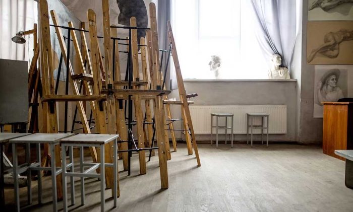 North Georgia Arts Guild Art studio with tall wooden easels stacked together, metal stools, and sketches on the wall near a bright window with curtains. - Clayton, GA