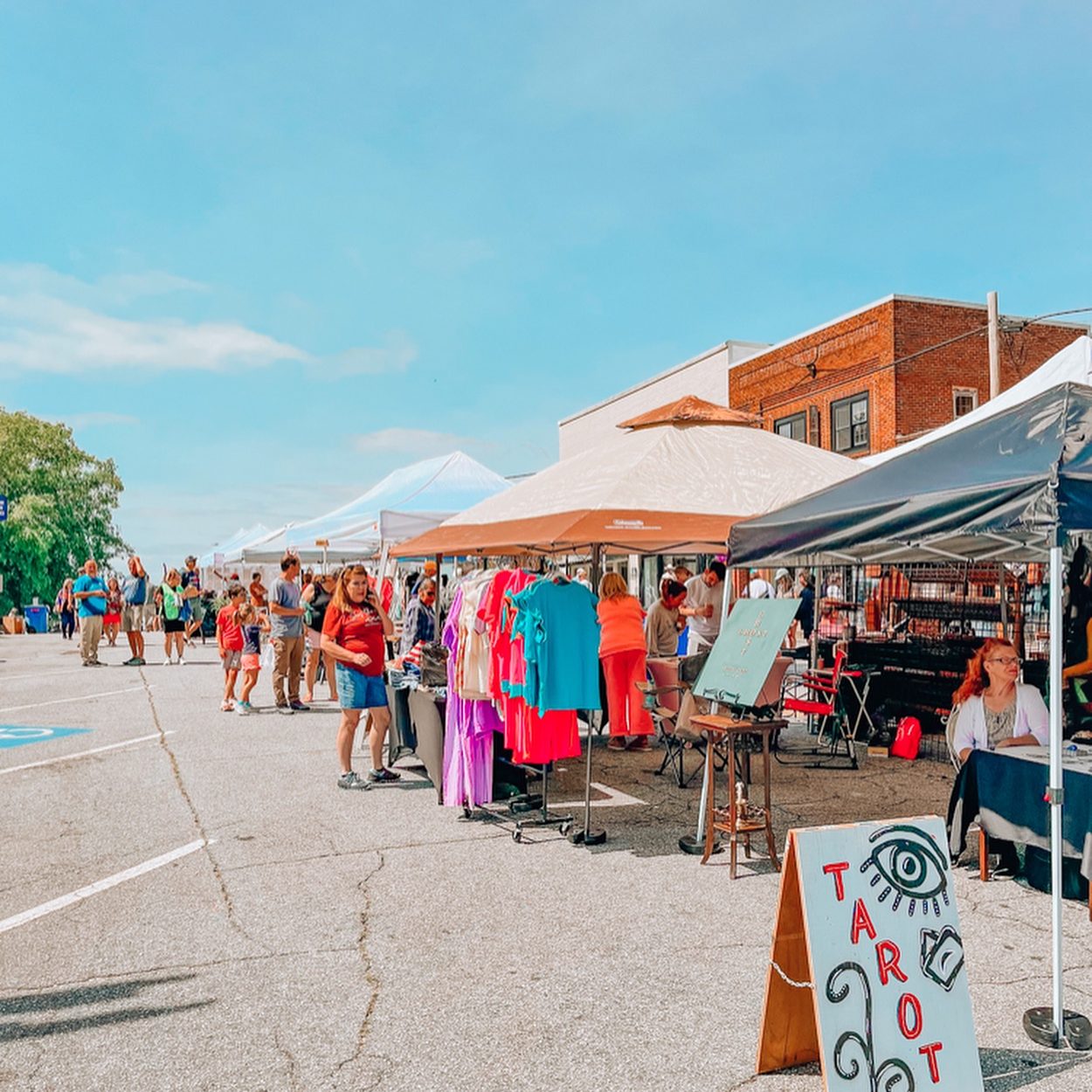 Outdoor street market with tents and shoppers browsing bright clothing on racks and tables under clear blue sky. - Clayton, GA
