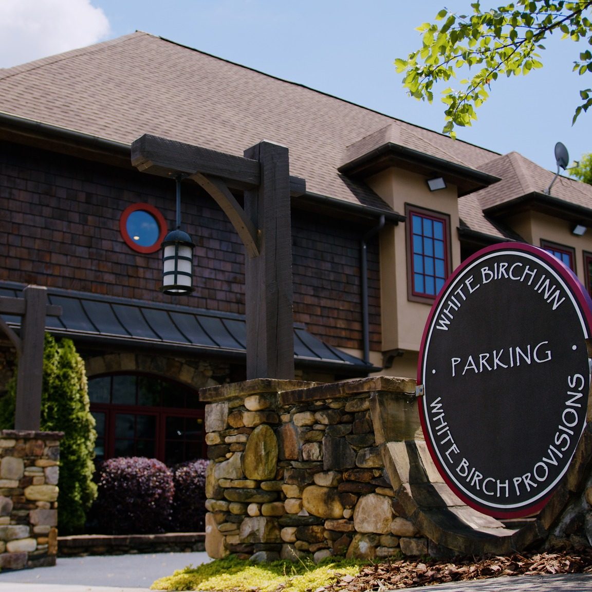 Exterior view of a rustic inn with brown shingles, stone wall, and hanging lanterns; a large circular sign reads 'White Birch Inn Parking' in the foreground. - Clayton, GA