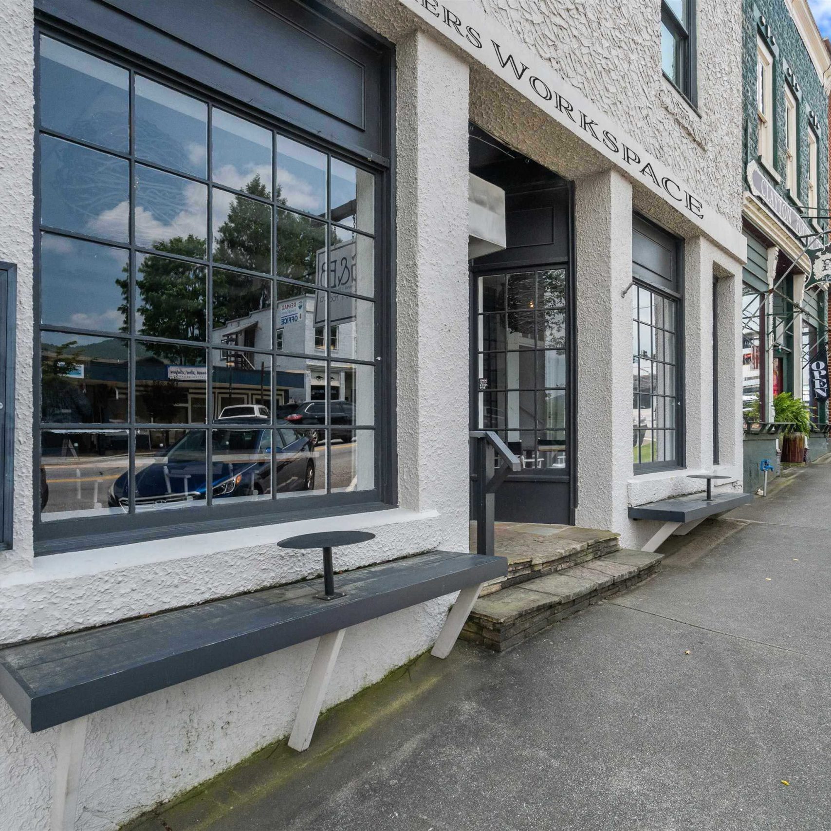 Row of storefronts along a sunny street with large grid windows, white textured walls, and a Clayton FlowerBed Gifts sign nearby indicating a flower shop. - Clayton, GA