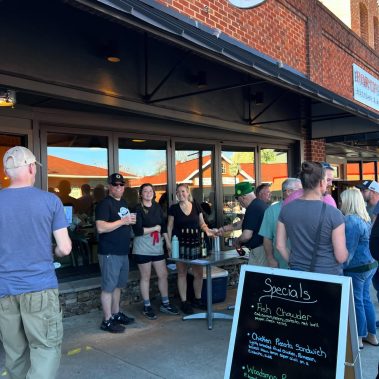 Group of people stand outside a brick storefront cafe, some holding drinks, next to a table with bottles and a chalkboard sign that says 'Specials' in front of the building (casual outdoor gathering). - Clayton, GA