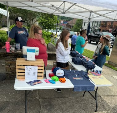 Vendor booth under a white canopy selling t-shirts, hats, and accessories; people browse and chat. - Clayton, GA