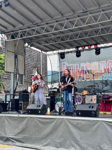 Female guitarist and vocalist performing on an outdoor stage with a drummer in the background. - Clayton, GA