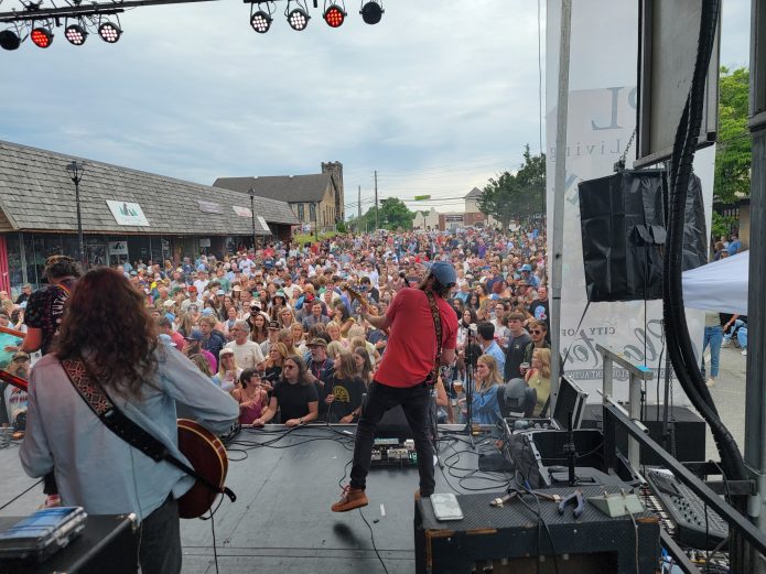Musician on stage performing to a large outdoor crowd; audience fills a street festival with buildings and trees in the background. - Clayton, GA