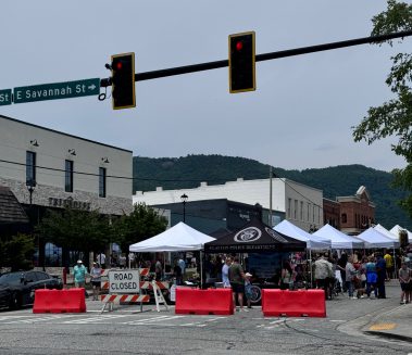 Street market on a closed street with white tents, shoppers, and orange barriers under a traffic light and East Savannah St sign behind them - Clayton, GA