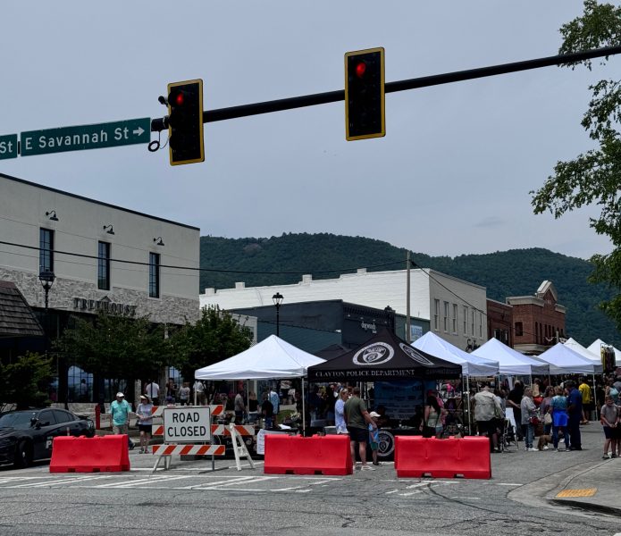 Street market on a closed street with white tents, shoppers, and orange barriers under a traffic light and East Savannah St sign behind them - Clayton, GA