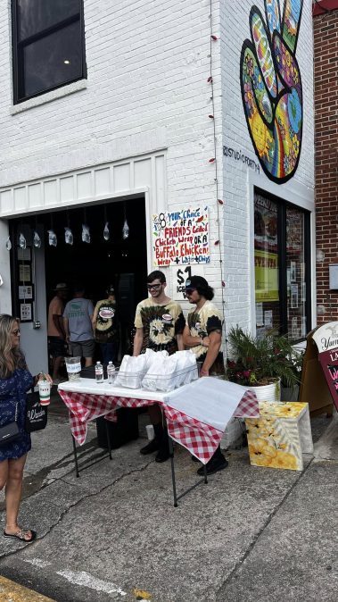 Outdoor street scene with a white brick building and colorful mural; two men in tie-dye shirts stand behind a table with a red-and-white checkered tablecloth handing out bags under a storefront menu sign. - Clayton, GA