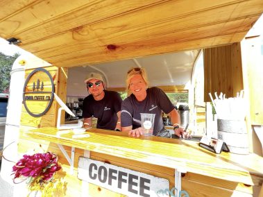 Two baristas in black work shirts lean from a wooden coffee cart with a 'COFFEE' sign and pink flowers in front. - Clayton, GA