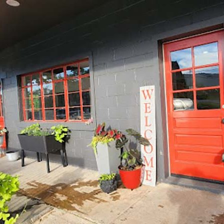 Red front door beside a gray brick wall, with large windows and potted plants along a concrete walkway. - Clayton, GA