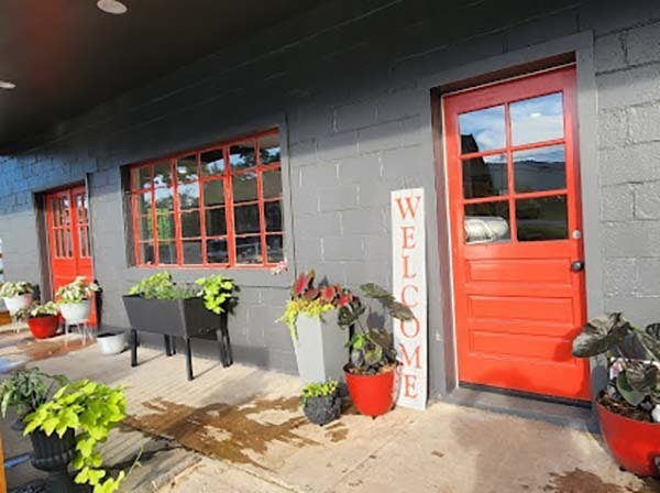 Rabun Flooring Red front door beside a gray brick wall, with large windows and potted plants along a concrete walkway. - Clayton, GA