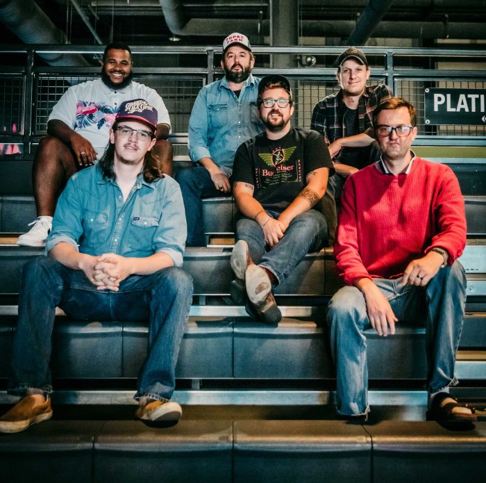 Six men sit on metal bleachers in an industrial setting, posing for a group photo in casual clothing. - Clayton, GA