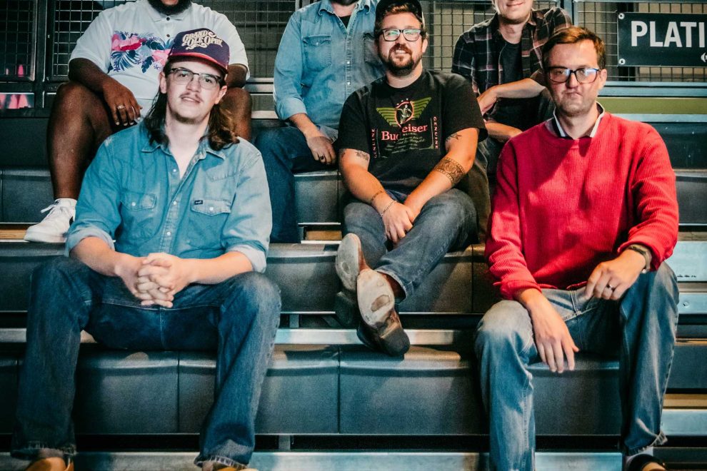 Six men sit on metal bleachers in an industrial setting, posing for a group photo in casual clothing. - Clayton, GA