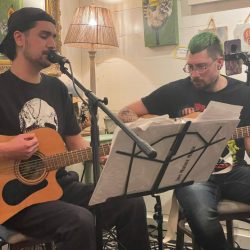Two men playing acoustic guitars and singing into microphones in a cozy, art-filled room with a lamp and artwork on the wall. - Clayton, GA