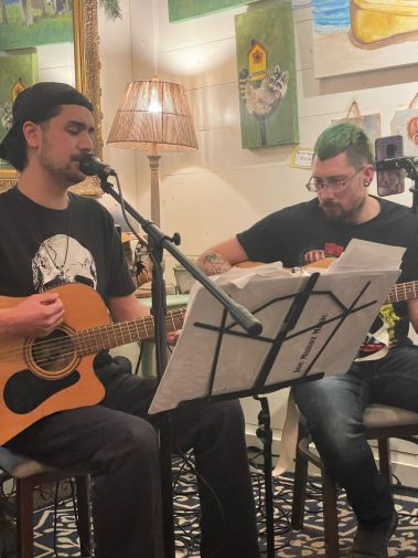 Two men playing acoustic guitars and singing into microphones in a cozy, art-filled room with a lamp and artwork on the wall. - Clayton, GA