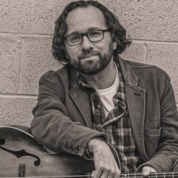 Portrait of a man with glasses and a beard, wearing a jacket and plaid shirt, leaning on an acoustic guitar against a brick wall. - Clayton, GA