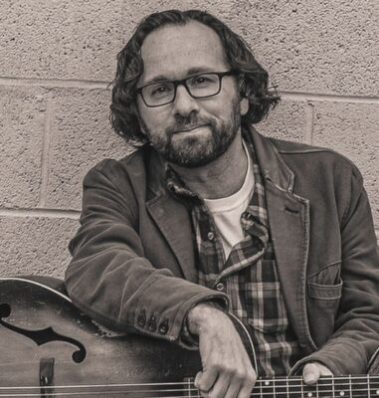 Portrait of a man with glasses and a beard, wearing a jacket and plaid shirt, leaning on an acoustic guitar against a brick wall. - Clayton, GA