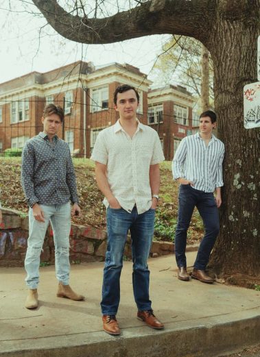 Three men stand on a sidewalk near a large tree in an urban park, posing for a photo in casual outfits. - Clayton, GA