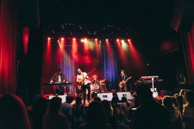 Live band performing on a stage bathed in red and yellow lights, with audience silhouettes in the foreground. - Clayton, GA