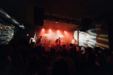 A live band performs on a dimly lit stage with red and white spotlights while a crowd watches closely from the foreground. - Clayton, GA