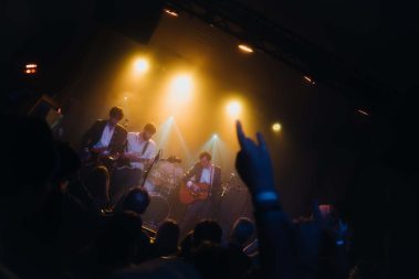 Band performs live on a dimly lit stage with warm yellow lights, guitarist center, drummer in back, and audience raising hands in front. - Clayton, GA