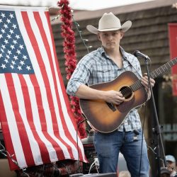 Male guitarist wearing a cowboy hat performing on an outdoor stage with a large American flag backdrop of red and white stripes and stars. - Clayton, GA