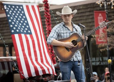 Male guitarist wearing a cowboy hat performing on an outdoor stage with a large American flag backdrop of red and white stripes and stars. - Clayton, GA