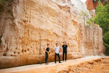 Three men stand on a dirt trail beside a tall orange rock cliff outdoors, wearing casual clothes and posing for the photo. - Clayton, GA