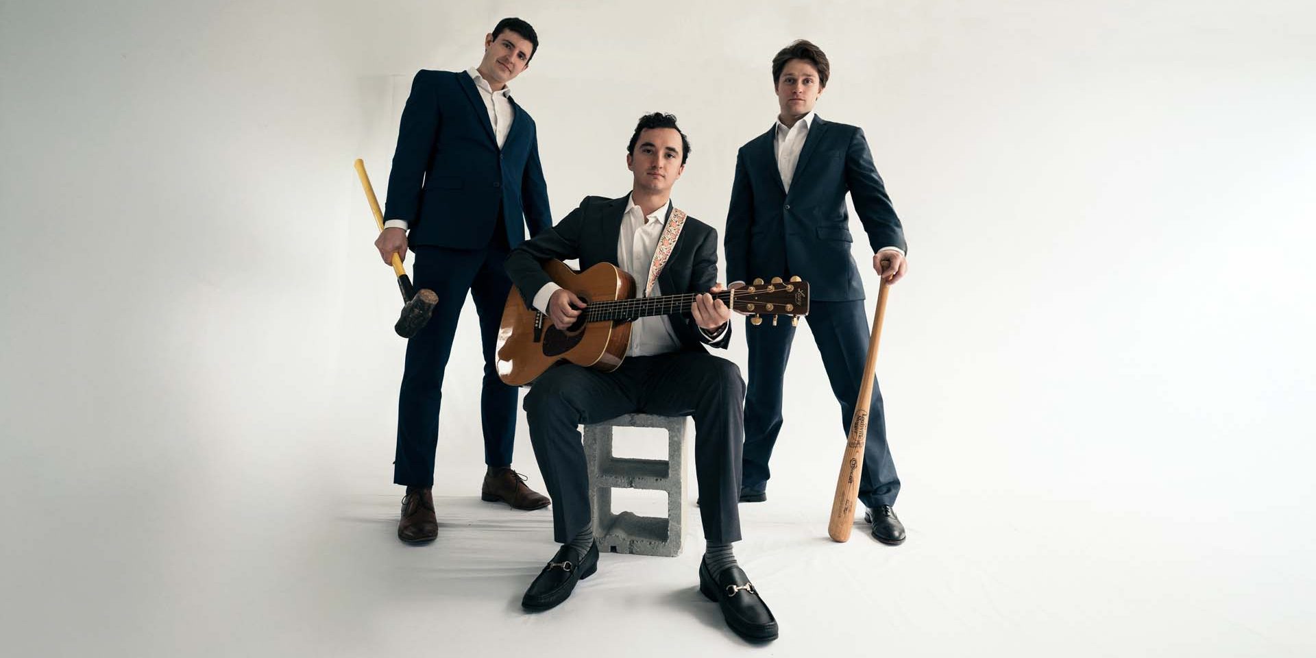 Kobanova Three men in suits with a guitarist seated on a concrete block, two others standing with wooden bats in a studio setting. - Clayton, GA