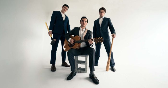 Three men in suits with a guitarist seated on a concrete block, two others standing with wooden bats in a studio setting. - Clayton, GA