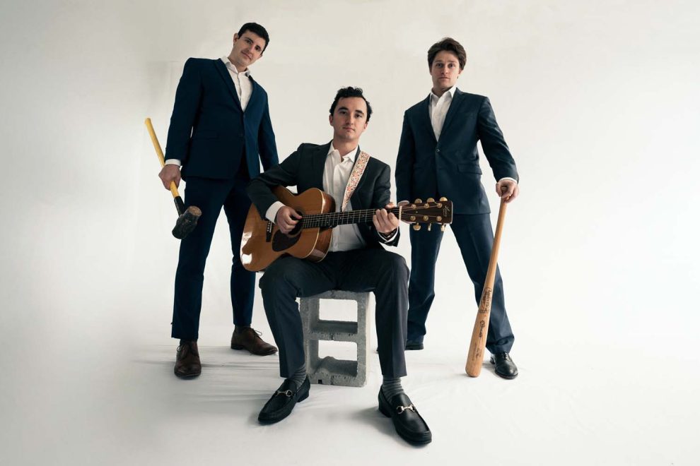 Three men in suits with a guitarist seated on a concrete block, two others standing with wooden bats in a studio setting. - Clayton, GA