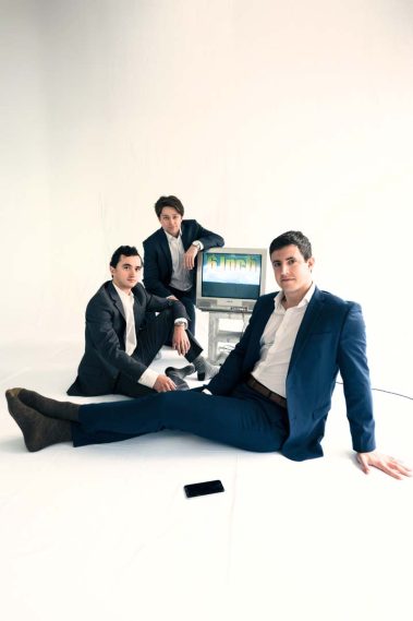 Three men in suits pose in a bright white studio; an old CRT monitor reads '6inch' and a smartphone lies on the floor. - Clayton, GA
