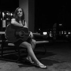 Young woman sits on a bench at night with an acoustic guitar resting on her lap under dim lights. - Clayton, GA