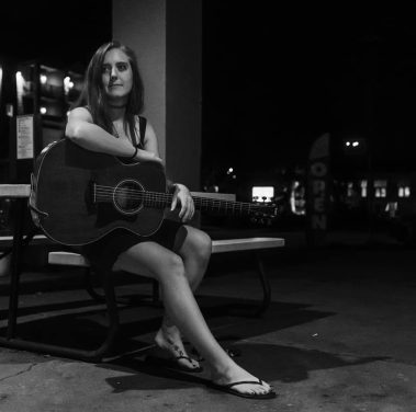 Young woman sits on a bench at night with an acoustic guitar resting on her lap under dim lights. - Clayton, GA