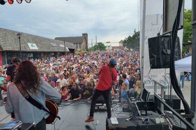 Live outdoor concert with a large crowd facing a stage; two guitarists perform, one in a red shirt center stage, other on the left with a guitar; stage gear and cables surround them. - Clayton, GA