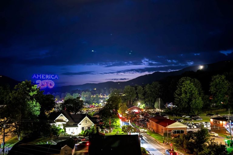 Nighttime hillside town with a blue illuminated sign reading 'AMERICA 50' on the left; cars and houses lit below. - Clayton, GA