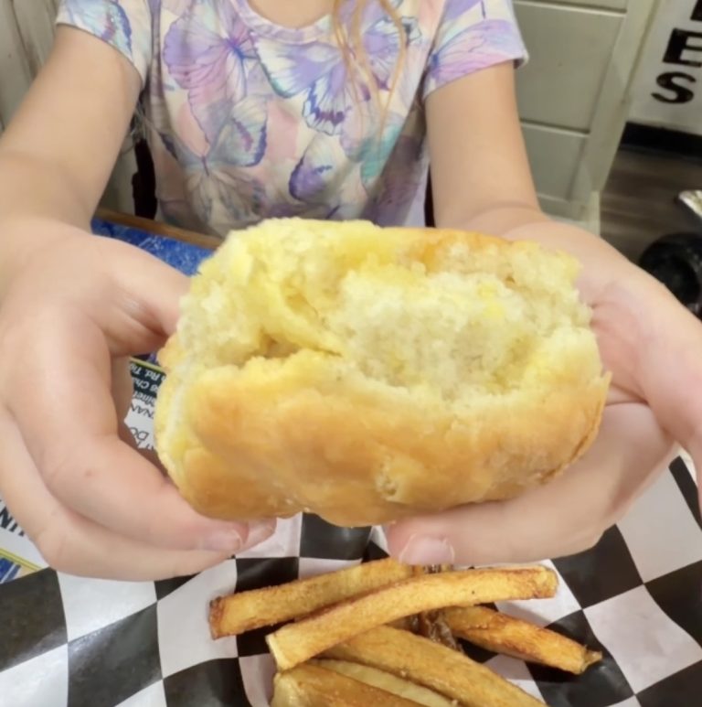 Person in a pastel tie-dye shirt holds a large hollow bread roll with a bite taken out, over a tray of fries on a checkered liner. - Clayton, GA