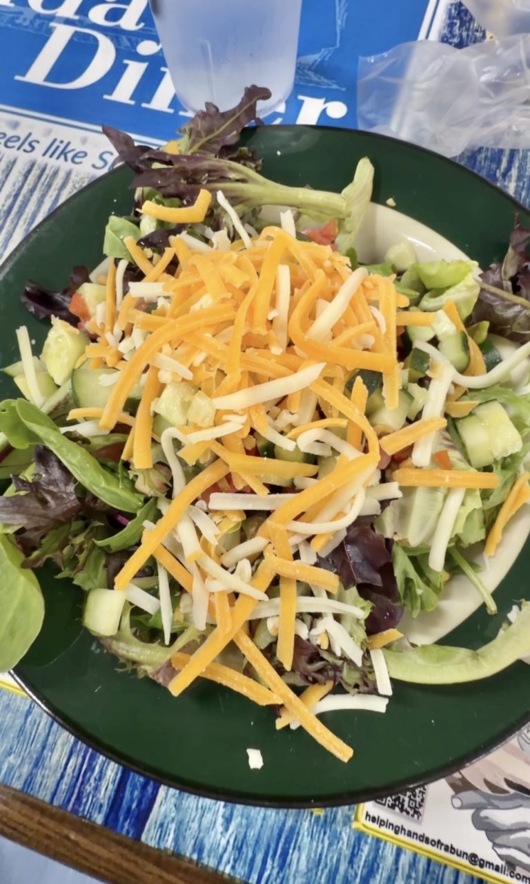 Green plate with a fresh garden salad topped with orange cheddar and white cheese shreds, plus cucumber and tomatoes on a blue table. - Clayton, GA