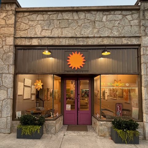 Storefront with purple double doors, bright orange sun logo above, stone facade, and planters on each side.